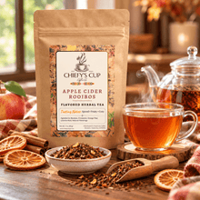 Load image into Gallery viewer, A kraft paper pouch of Chiefy's Cup Tea Apple Cider Rooibos flavored herbal tea stands on a wooden table, with a glass teapot, cup of amber tea, loose tea, dried orange slices, cinnamon sticks, and an apple in a warm, rustic kitchen setting.