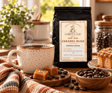 Load image into Gallery viewer, A black bag of Chiefy's Cup Caramel Bliss ground coffee stands on a wooden table beside a speckled mug of coffee, caramel pieces, coffee beans, and a slice of cake in a warm kitchen setting.