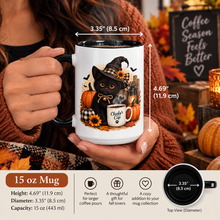 Load image into Gallery viewer, A person holds a 15 oz white ceramic mug with a black handle and rim. The mug features a fall-themed illustration of a black cat in a witch hat with pumpkins, sunflowers, and a coffee cup.