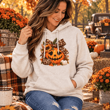 Load image into Gallery viewer, A woman wearing a light beige hoodie with a pumpkin jack-o-lantern graphic and leopard print bow, sitting in an autumn outdoor setting with pumpkins and fall foliage.