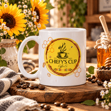 Load image into Gallery viewer, A white ceramic coffee mug with a yellow and orange circular graphic reading 'CHIEFY'S CUP Coffee Co' sits on a wooden board surrounded by coffee beans, sunflowers, a honey jar, and a cloth in a warm, rustic setting.