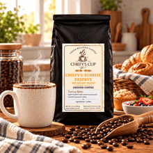 Load image into Gallery viewer, A black bag of Chiefy's Cup Coffee 'The Life' Chiefy's Sunrise Reserve Breakfast Blend ground coffee is displayed on a rustic wooden table. The scene is styled with a steaming ceramic mug, a wooden spoon of coffee beans, fresh croissants, and a jar of whole coffee beans in a warm, sunlit kitchen setting.