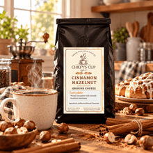 Load image into Gallery viewer, A bag of Chiefy's Cup Coffee Cinnamon Hazelnut Ground Coffee stands on a rustic wooden table in a warm, cozy kitchen setting. The scene includes a steaming mug of coffee, hazelnuts, cinnamon sticks, and a cinnamon roll in the background, creating an inviting atmosphere.