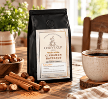 Load image into Gallery viewer, A black bag of Chiefy's Cup Cinnamon Hazelnut Ground Coffee stands on a wooden table, surrounded by hazelnuts, cinnamon sticks, a ceramic cup of coffee, and a softly blurred indoor background.