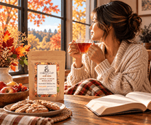 Load image into Gallery viewer, A cozy autumn lifestyle scene showing a woman in a cream knit sweater drinking tea at a wooden table, with an open book, cookies, and a stand-up pouch of Chiefy's Cup Apple Ciders Rooibos herbal tea in the foreground.