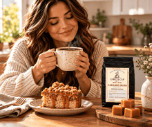 Load image into Gallery viewer, A woman in a cozy cream knit sweater holds a speckled mug with coffee, seated at a wooden table with a slice of caramel-topped cake, caramel candies, and a black bag of Chiefy's Cup Caramel Bliss Ground Coffee in a warm kitchen setting.