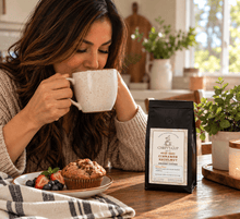 Load image into Gallery viewer, A woman in a cozy kitchen setting drinks from a speckled mug next to a bag of Chiefy's Cup Hazelnut Coffee. A muffin and berries sit on a plate on the table, with warm natural light coming from a window in the background.