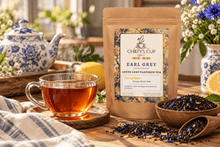 Load image into Gallery viewer, A kraft paper pouch of Chiefy's Cup Earl Grey Loose Leaf Flavored Tea stands on a wooden table beside a glass of amber tea, bowls of loose leaves, lemons, and floral decor in a warm, styled tea-room setting.