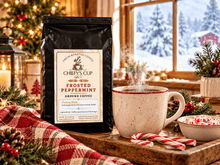 Load image into Gallery viewer, A bag of Chiefy's Cup Coffee Frosted Peppermint Ground Coffee stands on a wooden tray beside a steaming mug, candy canes, a small bowl of peppermint, and holiday greenery in front of a window showing a snowy outdoor scene.
