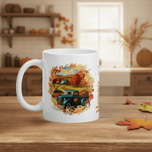 Load image into Gallery viewer, White mug with autumn-themed design featuring a blue truck and trees on a wooden table.
