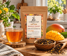 Load image into Gallery viewer, A stand-up pouch of Chiefy's Cup Loose Leaf Flavored Tea labeled Mango Treat stands on a wooden table, surrounded by a glass cup of amber tea, a bowl of loose tea leaves, a spoon of tea, and fresh mango slices and a whole mango in a bright kitchen setting.