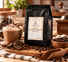 Load image into Gallery viewer, A black bag of Chiefy's Cup Midnight Mocha coffee stands on a wooden surface in a warm, styled coffee scene. A mug of hot chocolate with whipped cream and cocoa dusting, chocolate chunks, coffee beans, a wooden scoop, and a coffee grinder are arranged around it.