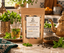 Load image into Gallery viewer, A stand-up kraft paper pouch of Chiefy's Cup Moroccan Mint Loose Leaf Tea stands on a marble countertop, surrounded by mint leaves, a glass of brewed tea, a wooden bowl of loose leaves, and a brass teapot in a warm kitchen setting.