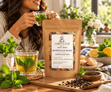 Load image into Gallery viewer, A stand-up kraft pouch of Chiefy's Cup Moroccan Mint Loose Leaf Tea stands on a wooden board in a bright kitchen scene. A woman in a cream sweater holds a glass of mint tea in the background, with mugs of tea, mint leaves, lemons, pastries, and loose tea visible around the product.