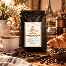 Load image into Gallery viewer, A black bag of Chiefy's Cup Coffee Parisian Ember French Roast Ground Coffee stands on a wooden table surrounded by coffee beans, chocolate pieces, a cup of coffee, croissants, and flowers in a warm, cozy cafe-like setting.
