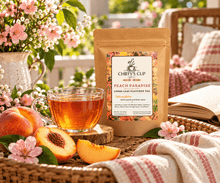 Load image into Gallery viewer, A stand-up pouch of Chiefy's Cup Peach Paradise loose leaf flavored tea stands on a woven tray beside a glass cup of amber tea, with sliced peaches, pink blossoms, an open book, and a soft blanket in a bright outdoor patio setting.