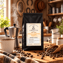 Load image into Gallery viewer, A black bag of Chiefy's Cup Chiefy's Frontier Reserve medium roast coffee stands on a wooden table in a warm, rustic kitchen setting. The scene includes a steaming moka pot, a ceramic mug, scattered coffee beans, and a wooden scoop.