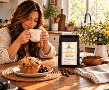 Load image into Gallery viewer, A woman in a cozy kitchen setting holds a speckled mug while looking at a bag of Chiefy's Barrel Reserve Medium Roast Whole Bean Coffee. A blueberry muffin sits on a plate in the foreground on a wooden table, with flowers and kitchen decor in the background.