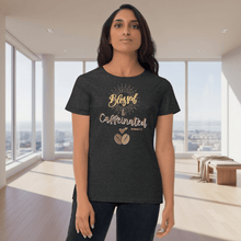 Load image into Gallery viewer, Front view of a woman wearing a dark gray “Blessed & Caffeinated” t-shirt with warm golden text and coffee bean art. The modern indoor background enhances the shirt’s cozy yet modern coffeehouse appeal.