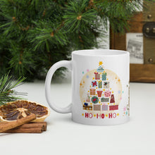 Load image into Gallery viewer, Left side view of a white glossy mug with an image design of a Christmas tree made of colorful presents, featuring a bright red image phrase "ho ho ho," with golden sparkles and a faded golden circle in the background.