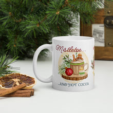 Load image into Gallery viewer, Left side view of a white glossy mug with the phrase "Mistletoe and Hot Cocoa" in festive font, featuring a design of a Christmas cup with a gingerbread man, whipped cream, ornaments, mistletoe, pine trimmings, and golden sparkles, with a round brown circle in the background.
