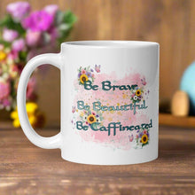 Load image into Gallery viewer, Left side view of a white glossy mug with the words &quot;Be Brave, Be Beautiful, Be Caffeinated&quot; displayed with floral arrangements on a pink and light blue hued background.
