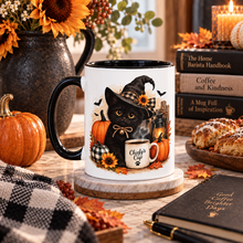 Load image into Gallery viewer, A white ceramic coffee mug with a black handle and interior, decorated with a black cat wearing a witch hat and surrounded by pumpkins, autumn leaves, and a small coffee cup. The mug is styled on a wooden coaster in a cozy fall-themed setting with books, a notebook, a candle, and a vase of sunflowers in the background.
