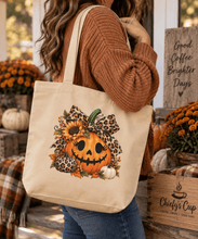 Load image into Gallery viewer, A woman wearing a brown ribbed sweater and blue jeans carries a beige canvas tote bag decorated with a jack-o'-lantern, leopard print bow, sunflowers, and autumn leaves. The scene is set in a cozy, decorated fall shop with pumpkins and a wooden sign reading 'Good Coffee Brighter Days'.

