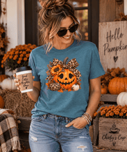 Load image into Gallery viewer, A woman wearing a heathered blue graphic t-shirt with a jack-o'-lantern, sunflowers, and leopard print design, holding a coffee cup in a fall-themed setting.
