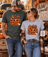 Load image into Gallery viewer, A man and a woman standing together in an autumn-themed outdoor setting, wearing matching graphic t-shirts. The man wears a dark green t-shirt and the woman wears a light gray t-shirt, both featuring a graphic of a jack-o'-lantern, sunflowers, leopard print, and a baseball.
