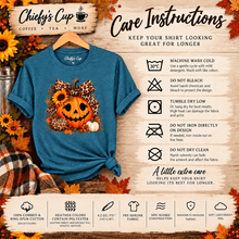 Load image into Gallery viewer, A blue graphic t-shirt with a fall-themed graphic print (leopard, cheetah, and white pumpkin) displayed on a wooden hanger over a plaid flannel shirt. The image is a care instructions infographic for 'Chiefsy's Cup Coffee, Tea, More' featuring various washing symbols and text overlays on a rustic wood background with autumn leaves.
