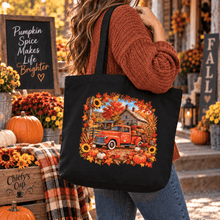 Load image into Gallery viewer, A woman wearing a rust-colored knit sweater and jeans carries a black tote bag with a colorful autumn-themed graphic of a red pickup truck surrounded by sunflowers and fall leaves. The background is a decorated fall porch with pumpkins, flowers, and a chalkboard sign.
