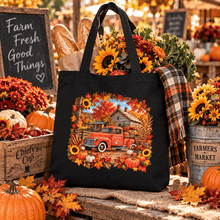 Load image into Gallery viewer, A black canvas tote bag with a large autumn-themed graphic print of a red pickup truck, pumpkins, sunflowers, and a barn, displayed in a rustic fall market setting with pumpkins, flowers, and a plaid scarf.
