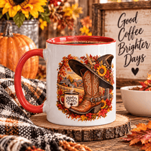 Load image into Gallery viewer, A white ceramic coffee mug with a red handle and rim, decorated with a western autumn scene featuring a cowboy boot, hat, fall leaves, and a small coffee cup logo. The mug is styled on a wooden slice with a plaid blanket, a pumpkin, and a sign reading 'Good Coffee Brighter Days'.
