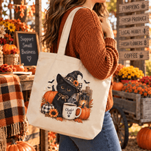 Load image into Gallery viewer, A woman wearing a rust-orange knit sweater and blue jeans carries a cream canvas tote bag with a Halloween-themed illustration of a black cat in a witch hat surrounded by pumpkins, a mug, and autumn flowers. The background is a rustic fall market stall with pumpkins, a plaid blanket, and wooden signs.
