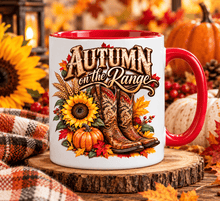 Load image into Gallery viewer, A white ceramic mug with a red handle and red interior, printed with the phrase 'Autumn on the Range' above a graphic of cowboy boots, pumpkins, sunflowers, and autumn leaves. The mug sits on a rustic wood slice surrounded by fall-themed props including pumpkins, sunflowers, berries, and a plaid blanket.
