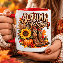 Load image into Gallery viewer, A person holds a white ceramic mug with a red handle and red interior. The mug features an autumn-themed graphic with the words 'AUTUMN on the Range,' cowboy boots, pumpkins, sunflowers, and fall leaves.
