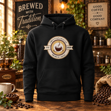 Load image into Gallery viewer, A black pullover hoodie displayed on a wooden table in a warm, rustic coffee-shop setting. The hoodie features a circular graphic on the chest with a coffee cup illustration and the text 'Cupcake Co.
