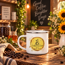 Load image into Gallery viewer, A white enamel coffee mug with a silver rim sits on a rustic wooden table in a cozy cafe-style setting. The mug features a circular yellow and green graphic with the text 'CHIEFY'S CUP COFFEE'.
