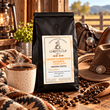 Load image into Gallery viewer, A black bag of Chiefy's Cup Coffee, Chiefy's Frontier Reserve Medium Bold Ground Coffee, styled on a rustic wooden table with a cup of coffee, scattered coffee beans, a wooden spoon, a cowboy hat, and a blanket in a warm, western-themed setting.
