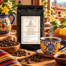 Load image into Gallery viewer, A bag of Chiefy's Cup Colombian Reserve ground coffee stands on a wooden table, surrounded by coffee beans, a colorful ceramic cup and saucer, flowers, and citrus fruits in a warm, sunlit setting.

