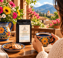 Load image into Gallery viewer, A person at a wooden table holds a colorful floral ceramic mug while a black bag of Chiefy's Cup Colombian Coffee stands beside a plate of cookies. Bright flowers in a matching ceramic pitcher and a scenic mountain village background create a warm, inviting atmosphere.
