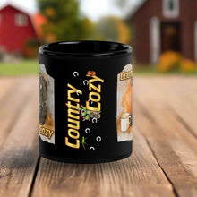 Load image into Gallery viewer, Back side view of a black glossy mug showcasing the phrase &quot;Country Cozy&quot; with a design of a black cowboy hat, flowering cactus, small lizard, and horse tracks.
