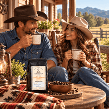 Load image into Gallery viewer, A man and woman in cowboy hats sit on a rustic wooden porch, each holding a mug, with a bag of Chiefy's Cup Coffee on the table between them. The setting includes a lantern, a bowl of coffee beans, and a mountainous outdoor background.
