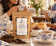 Load image into Gallery viewer, A stand-up pouch of Chiefy's Cup English Breakfast Loose Leaf Tea is centered on a breakfast table, surrounded by teacups, scones, jam, and fruit. Two people are partially visible in a bright, cozy kitchen setting, holding matching blue-and-white patterned cups.

