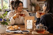 Load image into Gallery viewer, Two women sitting at a wooden table in a cozy kitchen setting, holding glass cups of tea. In the foreground, a stand-up pouch of Earl Grey tea by Chiefy's Cup is displayed alongside a glass teapot, a small bowl of loose leaves, and scones.
