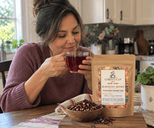Load image into Gallery viewer, A woman in a cozy kitchen setting holds a glass of dark herbal tea next to a stand-up pouch of Chiefy's Cup Hibiscus Berry Flavored Herbal Tea. A small wooden bowl filled with loose tea ingredients sits on the table in front of the package.

