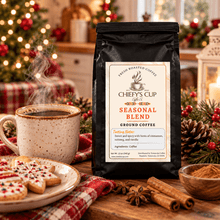 Load image into Gallery viewer, A black bag of Chiefy's Cup Seasonal Blend Ground Coffee stands on a wooden table in a cozy holiday setting, surrounded by a steaming mug, cookies, cinnamon sticks, and a blurred Christmas tree in the background.
