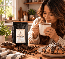 Load image into Gallery viewer, A woman in a cozy kitchen setting holds a white speckled mug, smiling at a bag of Chiefy's Cup Midnight Mocha coffee. The table is styled with chocolate pieces, coffee beans, a plate with a chocolate-dripped pastry, and a checkered blanket.

