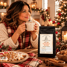 Load image into Gallery viewer, A woman in a cozy knit sweater and red plaid scarf holds a speckled mug in a warm holiday kitchen scene. A black bag of Chiefy's Cup Seasonal Blend ground coffee stands on the table beside cookies, cinnamon sticks, and a lit lantern, with a blurred Christmas tree in the background.
